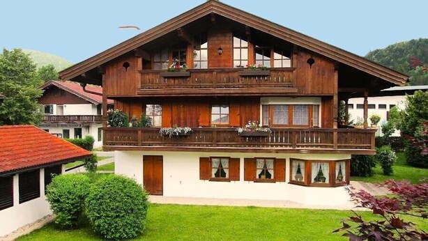 Un chalet en bois à deux étages, doté de balcons, d’une façade blanche et entouré d’un jardin.