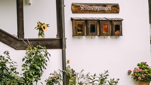 Un edificio rustico con facciata in legno, un muro bianco con un cartello che riporta la scritta "Fleischkuchen" e un piccolo giardino con piante e fiori.