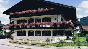 A two-story building with balconies adorned with flowers, a visible entrance, and a street view. - Ein-Raum-Ferienwohnung (3) 27qm, Bad/WC, Küchenzeile, Balkon (Reit im Winkl)
