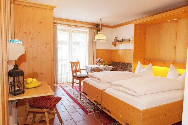 A wooden room with two beds, a table with a lamp, a chair, and a window with curtains. - Ein-Raum-Ferienwohnung (3) 27qm, Bad/WC, Küchenzeile, Balkon (Reit im Winkl)