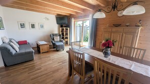 Uma sala de estar aconchegante com sofá, poltrona, mesa pequena e mesa de jantar com cadeiras. Teto de madeira e janela com cortinas.