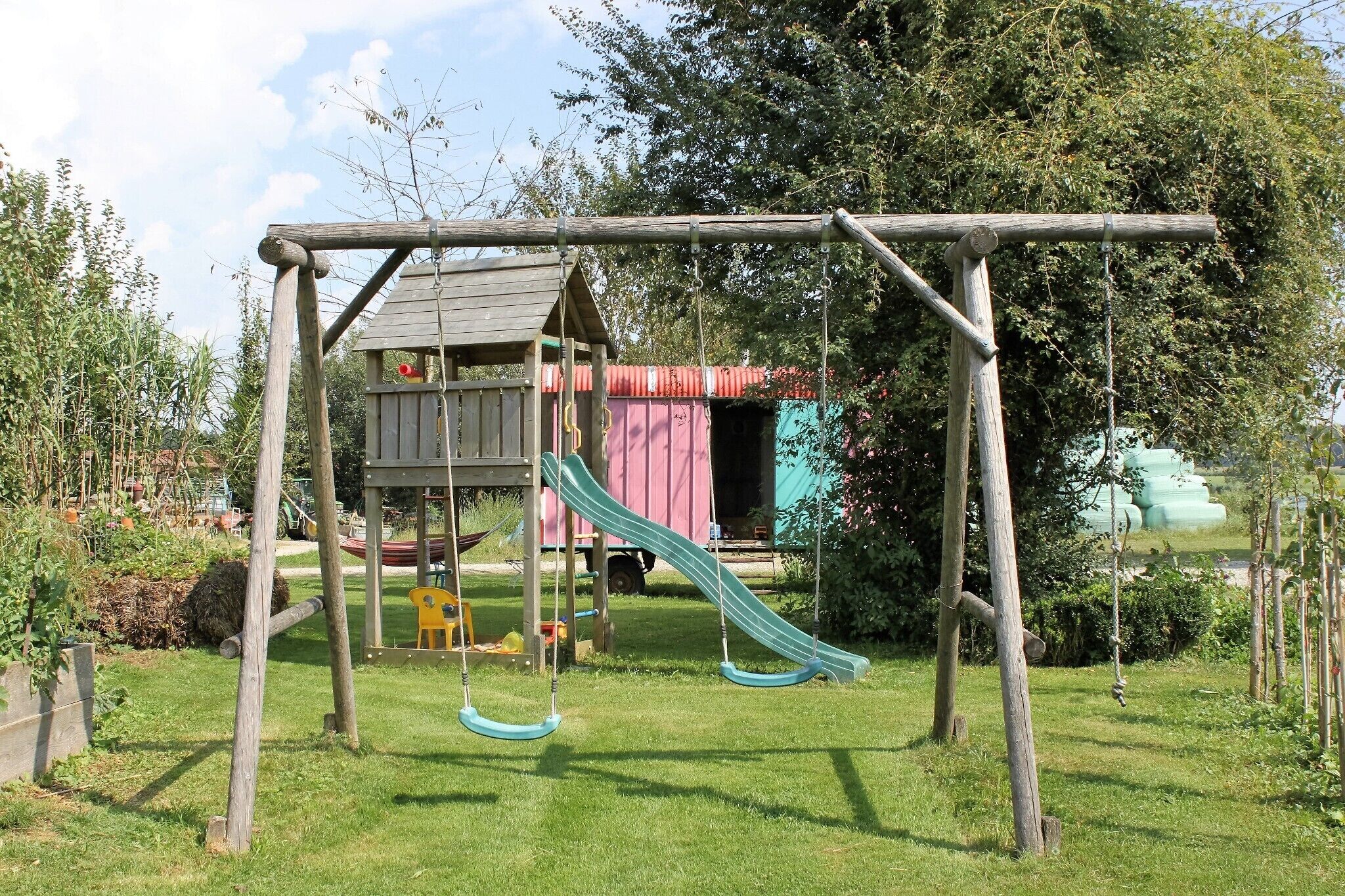 A wooden swing set with a slide, a small wooden house, and a truck in the background.