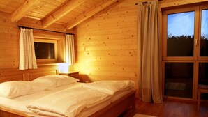 Ein Schlafzimmer einer Holzhütte mit einem Bett, Nachttischen, einem Fenster mit Vorhängen und einer Schiebetür zum Garten.