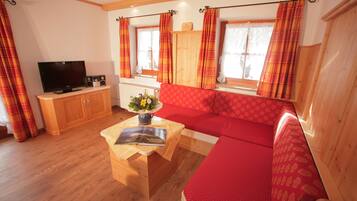 Ein gemütliches Wohnzimmer mit einem roten Sofa, einem Holz-TV-Schrank und einem kleinen Tisch mit Buch und Blumen.