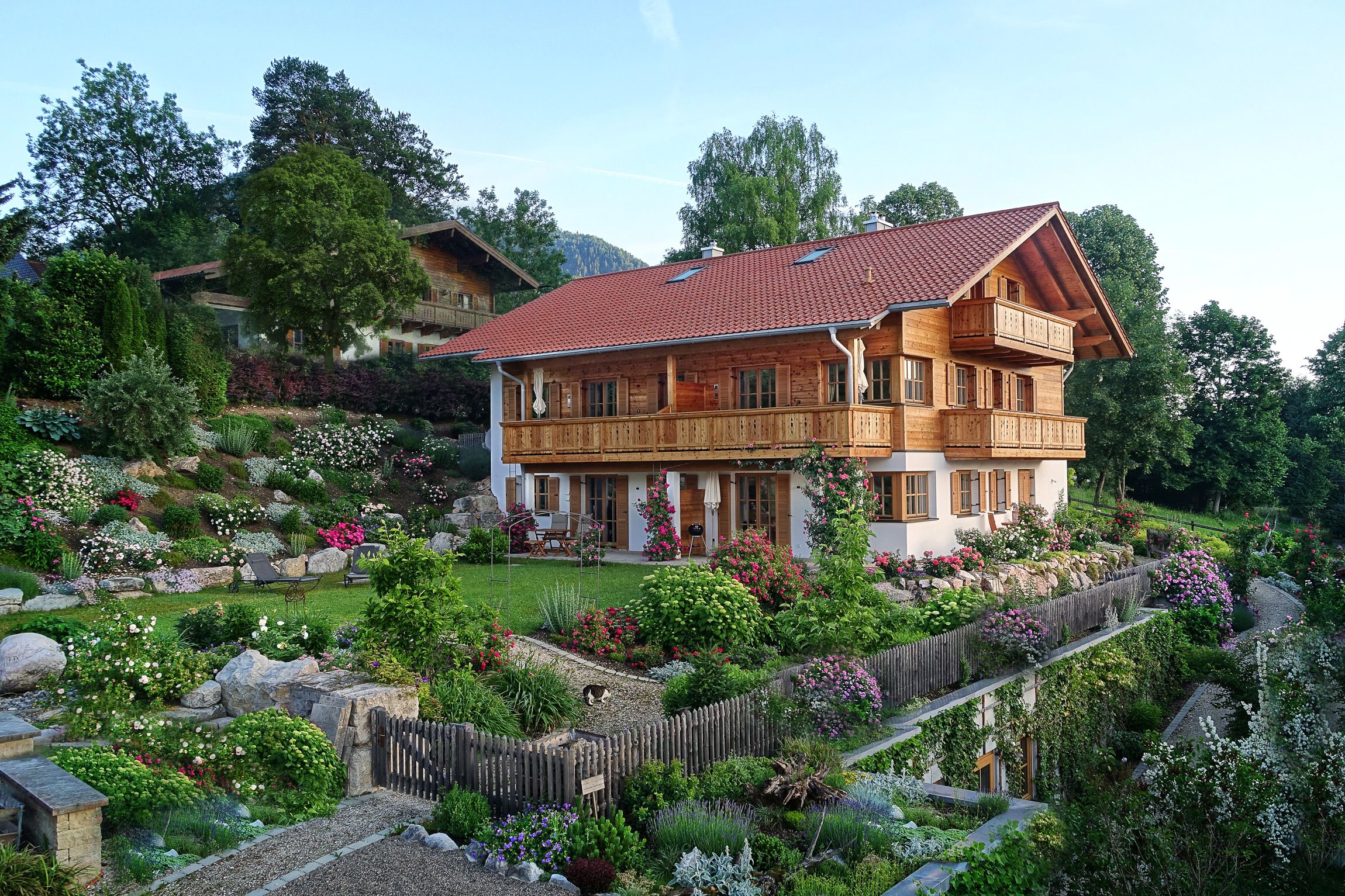Ein zweigeschossiges Holzhaus mit rotem Ziegeldach, umgeben von einem gepflegten Garten mit verschiedenen Pflanzen und Blumen.