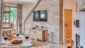 Un coin repas avec une table en bois dressée pour le repas, un meuble en bois sur lequel est posé un téléviseur à écran plat, et un mur en pierre.