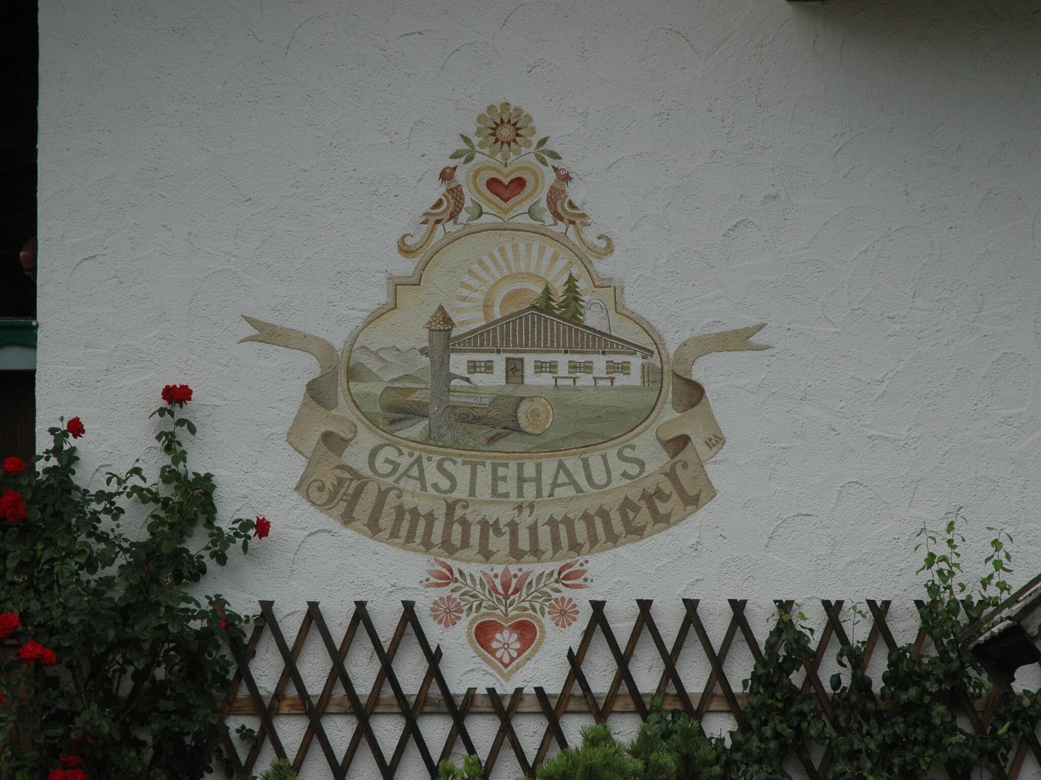 A wall with a logo for Gastehaus Almbrunner featuring a building, trees, and a sun.