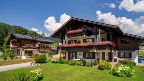 Un bâtiment en bois à deux étages, doté de balcons ornés de fleurs, entouré de jardins bien entretenus et surplombé d’un ciel dégagé.