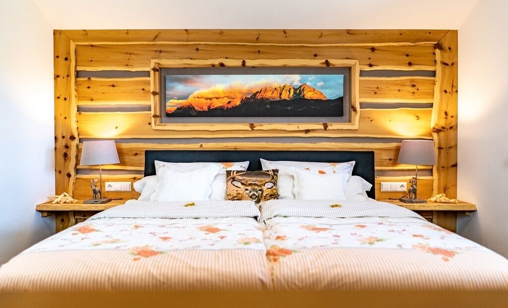 A double bed with floral bedding, two bedside lamps, a wooden headboard with a framed picture, and a wooden wall paneling.