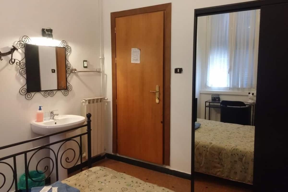 A hotel room with a wooden door, a sink, a mirror, a radiator, a bed with a floral bedspread, and a window with curtains.