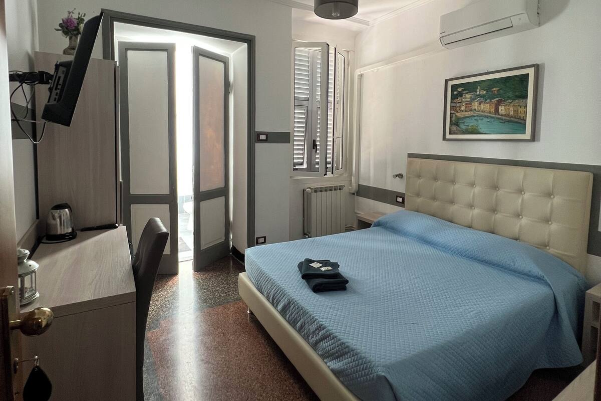 A hotel room with a bed, a desk, a chair, a radiator, and a painting on the wall.