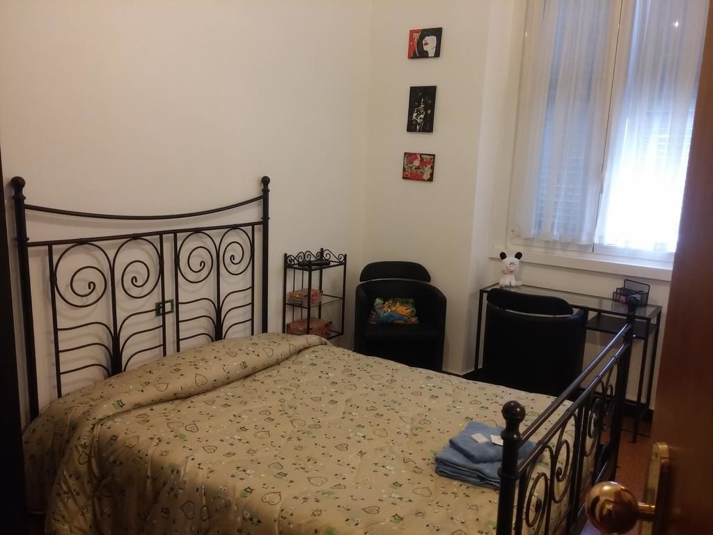 A bedroom with a metal bed frame, a patterned bedspread, a small bedside table, a chair, and a window with sheer curtains.