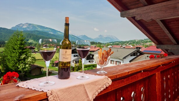 A bottle of wine and two glasses on a wooden balcony with a view of mountains and houses. - Ferienwohnung (9) (Reit im Winkl)