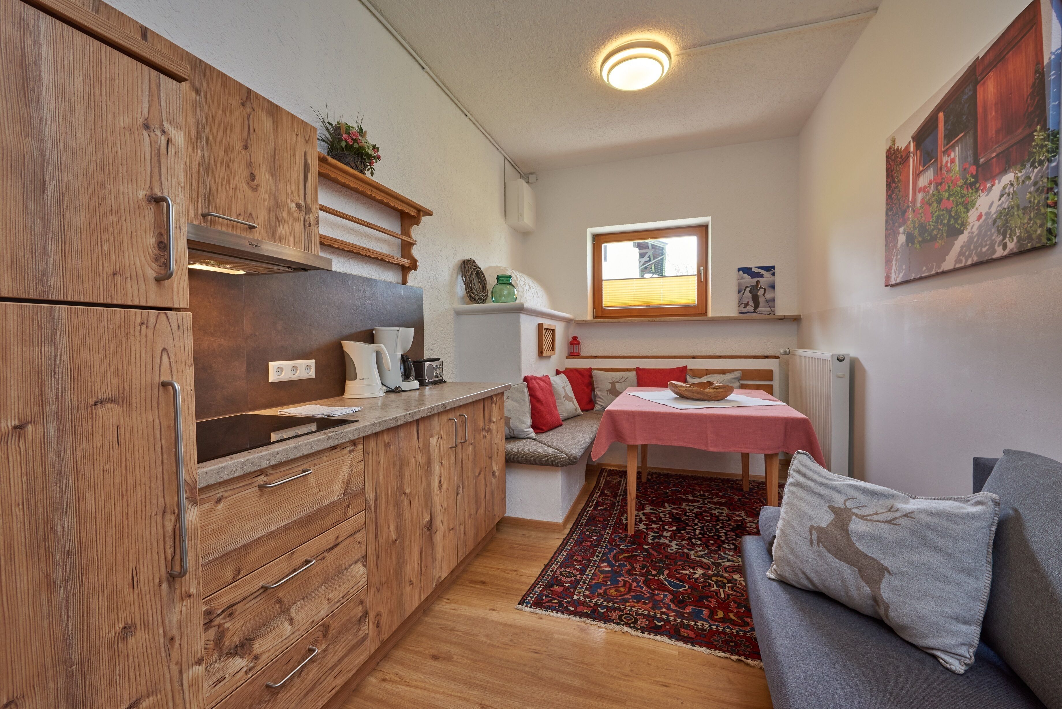 Ett kompakt kök med träskåp, en matplats med ett rött bord och en soffa med en kudde med ett hjortmotiv.