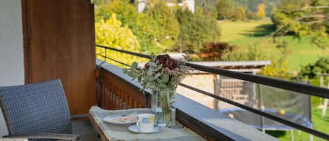 A balcony with a table set for two, a vase with flowers, and a view of a green landscape with trees and a building in the distance.