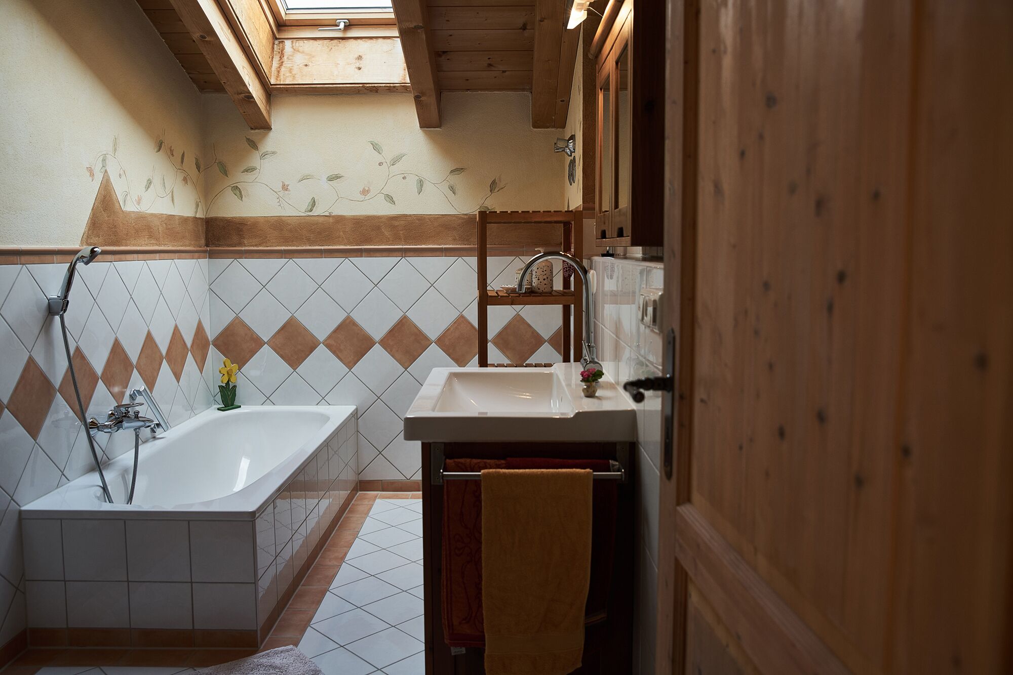 Une salle de bain avec une baignoire, un lavabo et une douche. Un puits de lumière se trouve au-dessus de la baignoire, et les murs sont recouverts de carreaux disposés en motif losange.