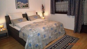 A bedroom with a bed, a nightstand, a lamp, a window with curtains, and a wall-mounted picture. - Apartment Rauschbergblick 80 sqm, 2 separate bedrooms, bathroom, eat-in kitchen (Ruhpolding)