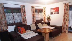 A living room with a dark brown sofa, a wooden coffee table, and floral curtains. - Apartment Rauschbergblick 80 sqm, 2 separate bedrooms, bathroom, eat-in kitchen (Ruhpolding)