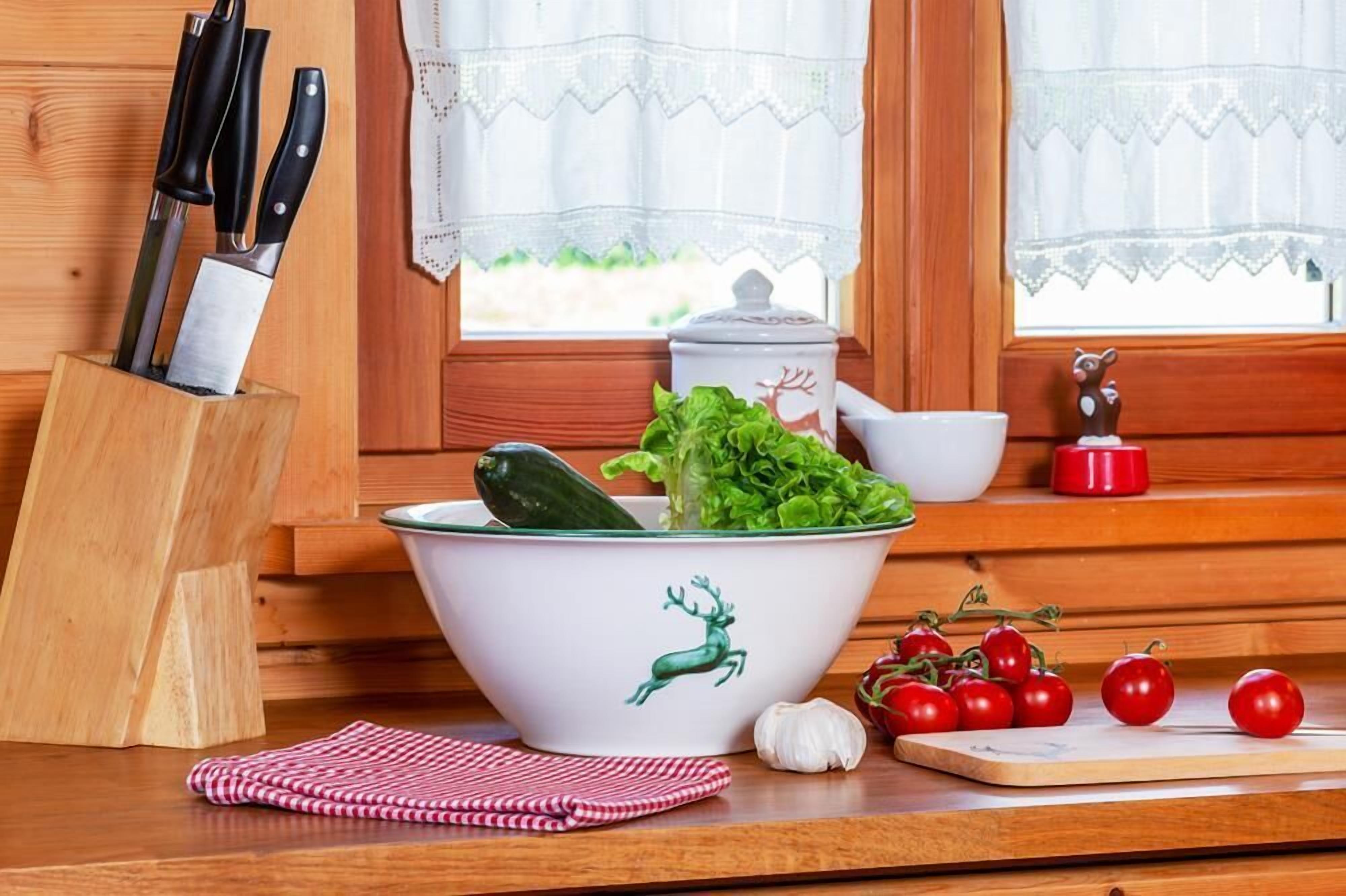 Une cuisine avec un plan de travail en bois, un saladier orné de motifs de rennes, un range-couteaux, une planche à découper avec des tomates et une fenêtre avec des rideaux en dentelle.