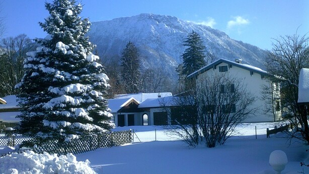 Un paysage enneigé avec une montagne en arrière-plan, une maison à l’extérieur blanc et au toit foncé, et un arbre recouvert de neige au premier plan.