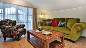 A living room with a green sofa, floral armchair, wooden coffee table, and a large window with sheer curtains.