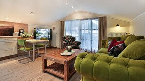 A modern living room with a green sofa, a wooden coffee table, and a floral armchair. There is a television and a dining area with a table and chairs.