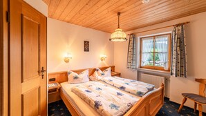Un dormitorio con techo de madera, una cama con ropa de cama floral, dos lámparas de noche, una ventana con cortinas y un radiador.