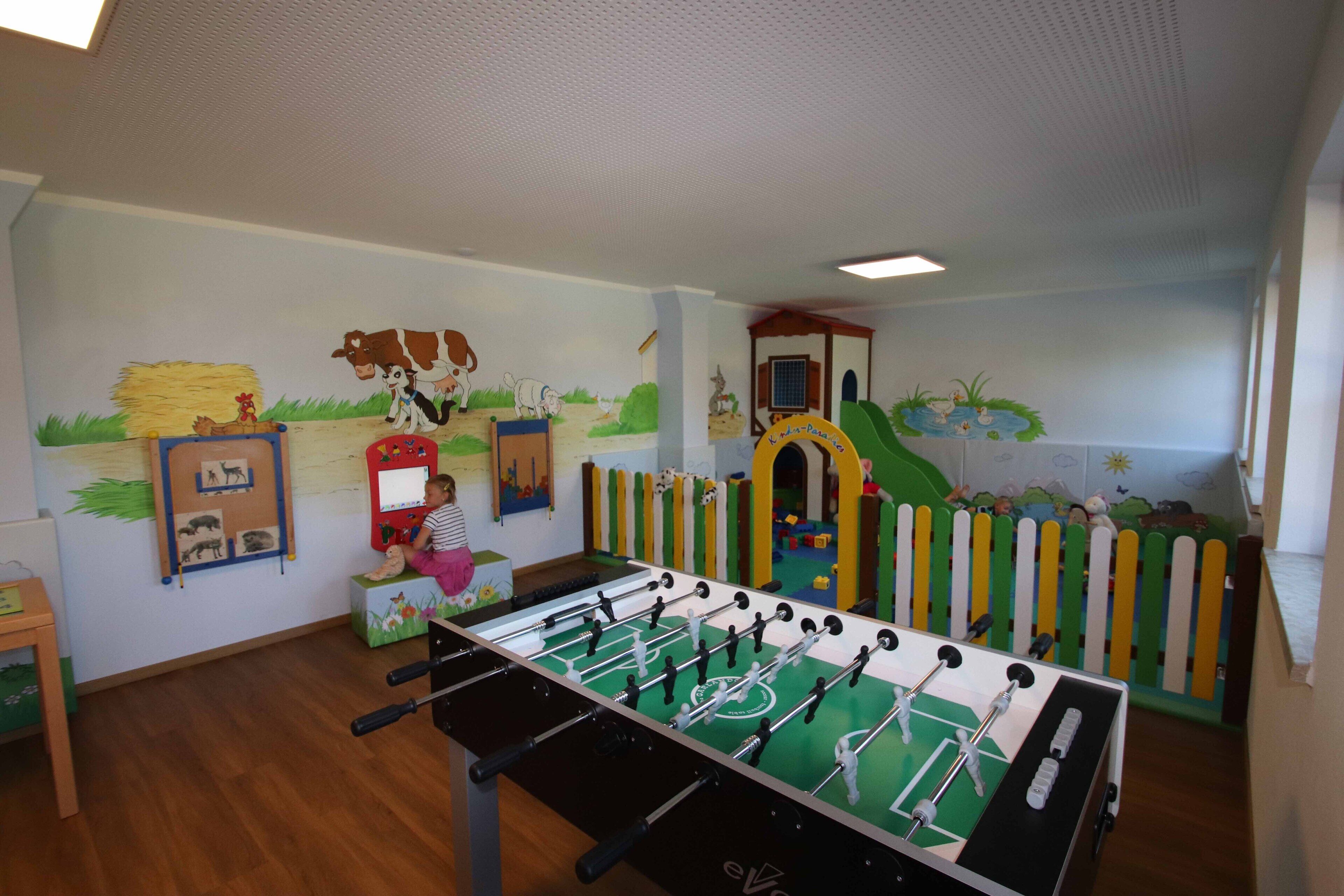 Una habitació amb una taula de futbolí, una zona de jocs amb tobogan i un mural amb animals de granja.