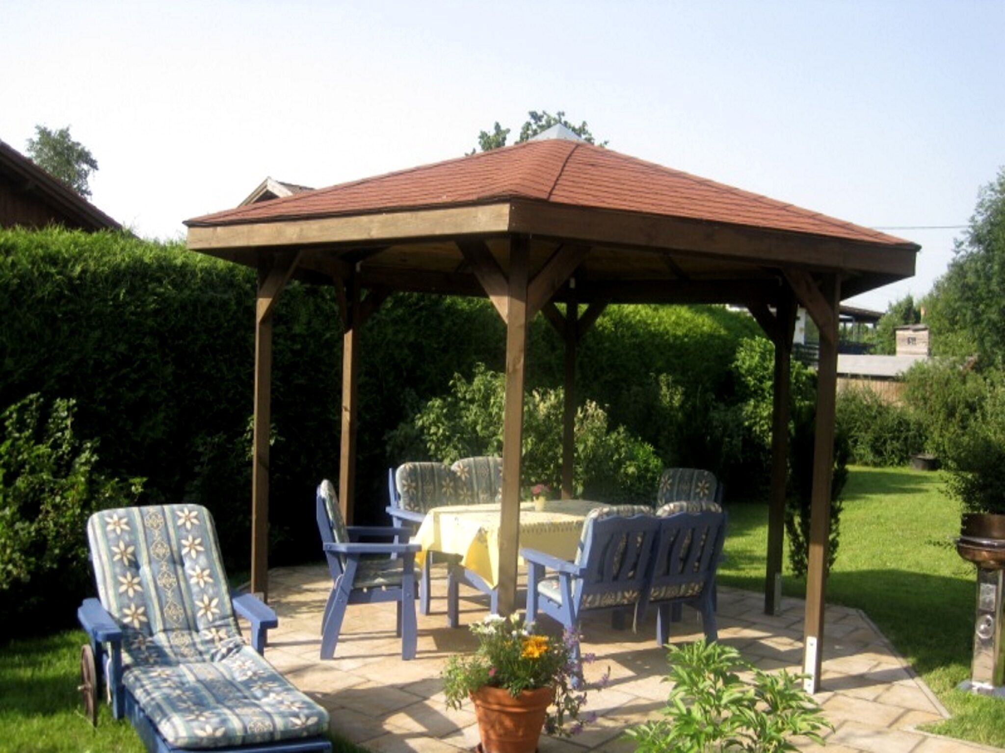 A covered patio area with a table and chairs, surrounded by greenery.