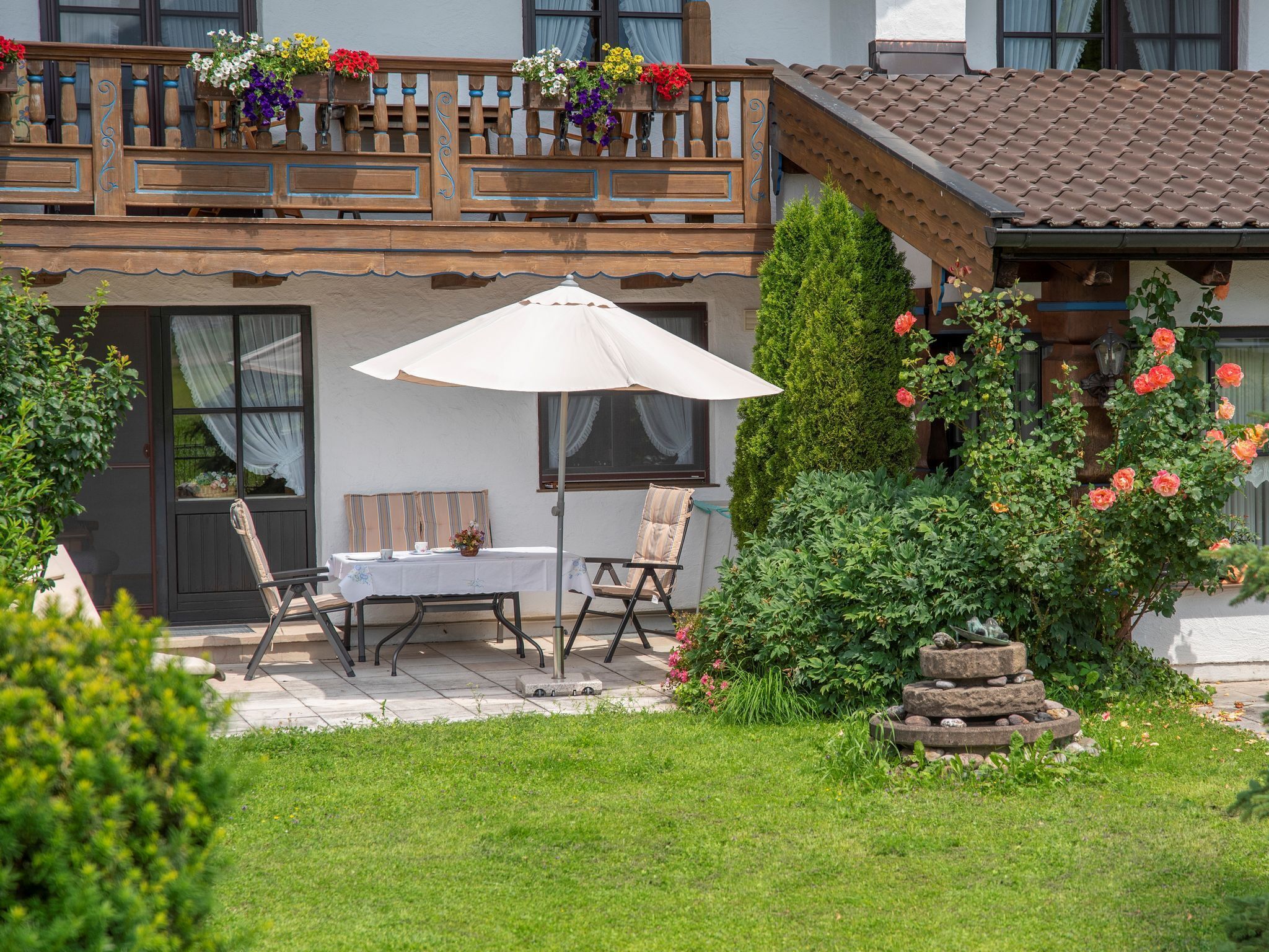 En terrasse med et bord og stole, en hvid parasol, samt en have med blomster og buske.