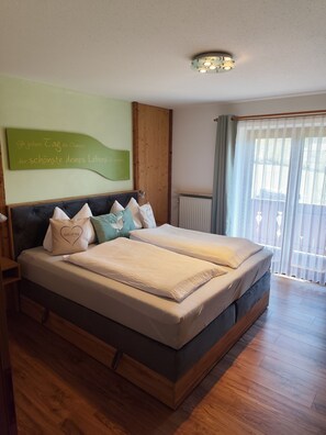 A double bed with a wooden frame, a headboard, and multiple pillows. There is a wall-mounted light fixture above the bed and a radiator on the wall. - Ferienwohnung Halsalm, 1-2 Personen, 43 qm, Dusche/WC, Balkon, TV, W-LAN, 1 Schlafzimmer, Nichtraucher, Süd-West-Lage (Ramsau bei Berchtesgaden)
