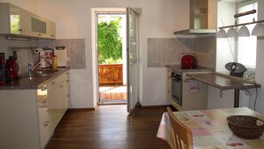 Moderni keittiö, jossa on puulattia, ruokailutila pöytineen ja tuoleineen sekä näkymä puukannelle avonaisen oven kautta.