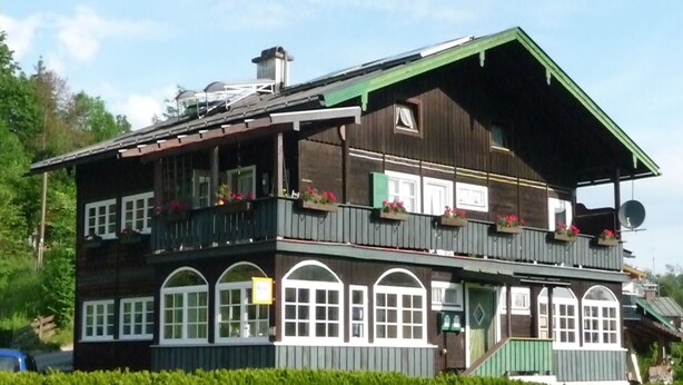 A two-story building with a green roof, multiple balconies with flower boxes, and a sign indicating a restaurant or cafe. - Ferienwohnung 65 qm, für 2-4 Personen, 2 getrennte Schlafzimmer, Balkon u. Nichtraucher (Schönau am Königssee)