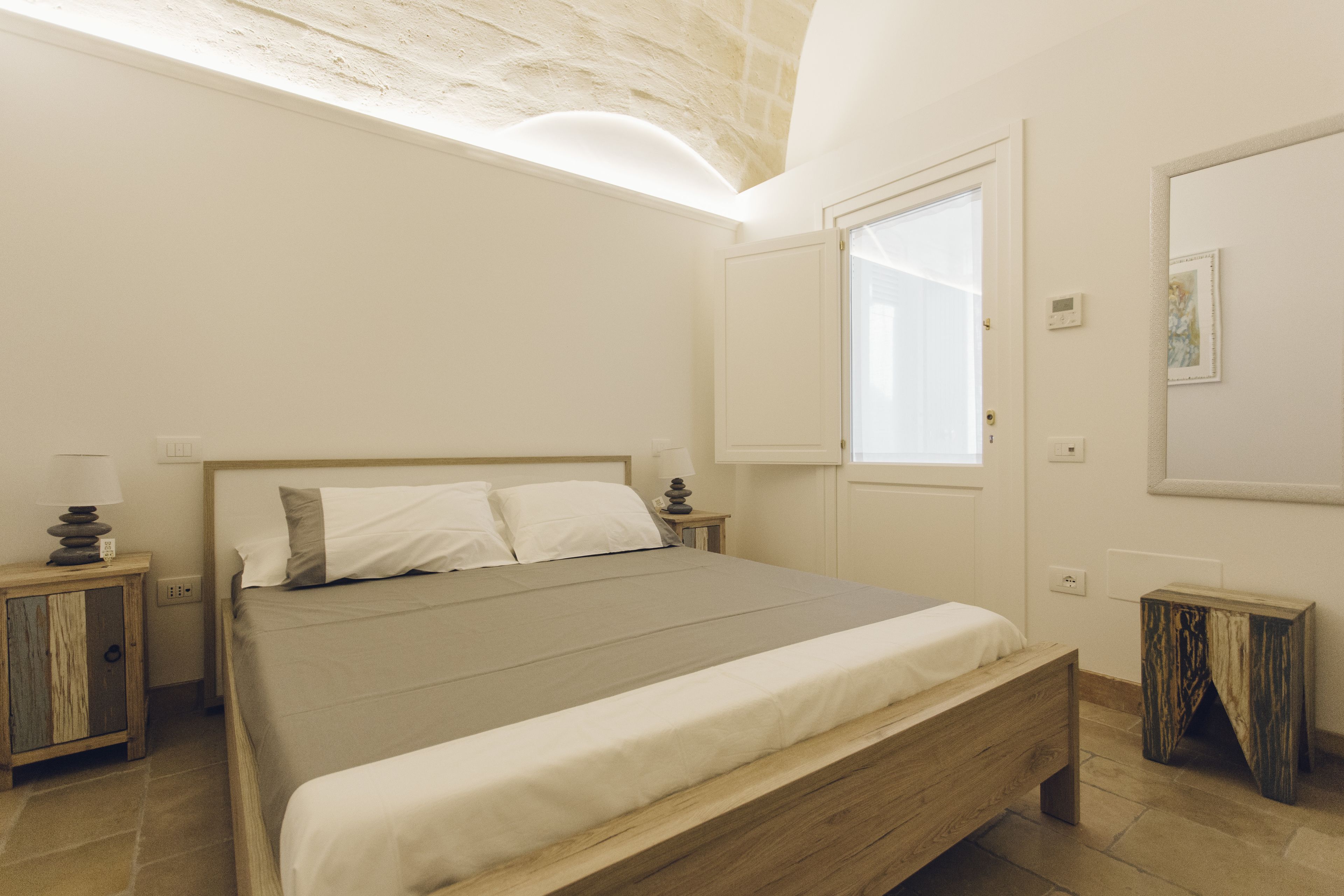 Une chambre à coucher avec un lit en bois, une table de chevet, un petit tabouret en bois, une fenêtre et un luminaire fixé au mur.