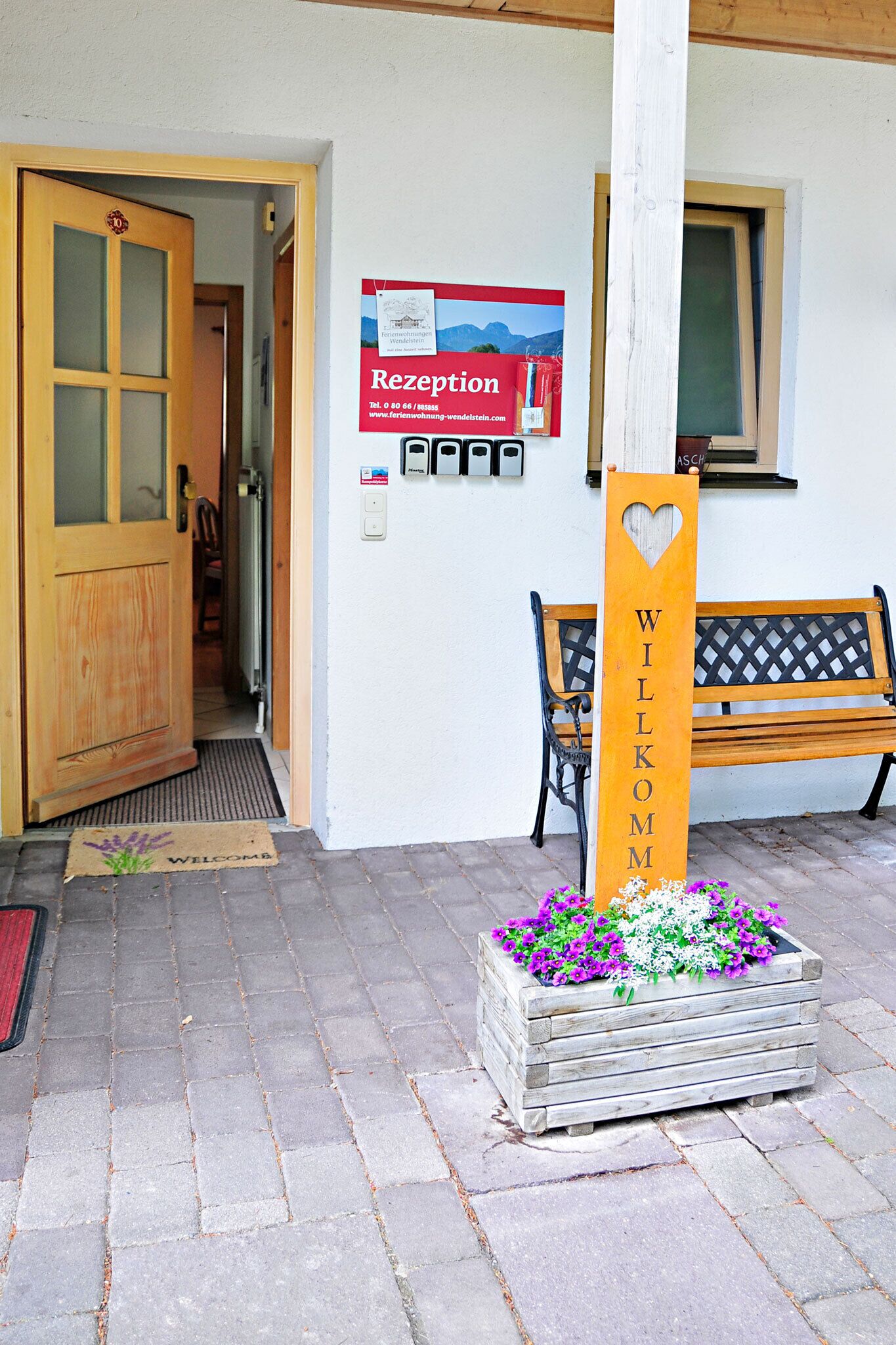 Un área de recepción con una puerta de madera, un banco, un felpudo, un parterre de flores y un letrero con un corazón recortado.