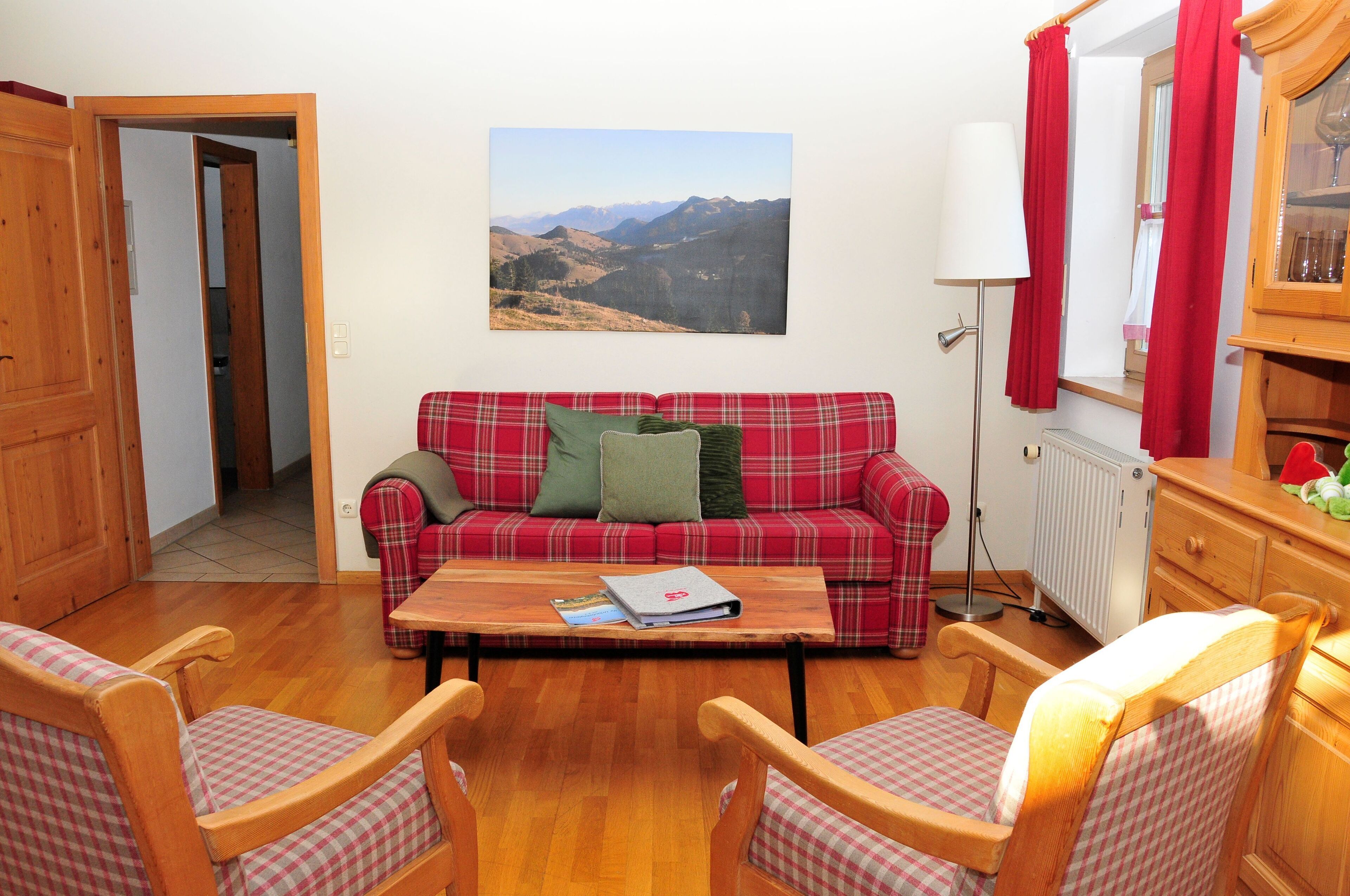 A cozy living room with a plaid sofa, wooden chairs, a coffee table with books, a view of mountains, and a red curtain.