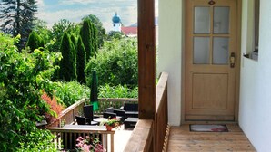 Une terrasse en bois avec une porte donnant sur un jardin, le tout entouré de verdure et sous un ciel dégagé.