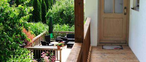 Une terrasse en bois avec une porte donnant sur un jardin, le tout entouré de verdure et sous un ciel dégagé.