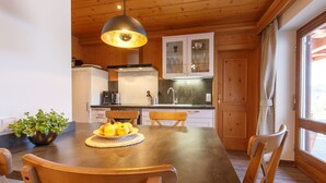 Una mesa de comedor de madera con sillas, un cuenco de fruta y una lámpara colgante en una cocina con armarios de madera y un ventanal.