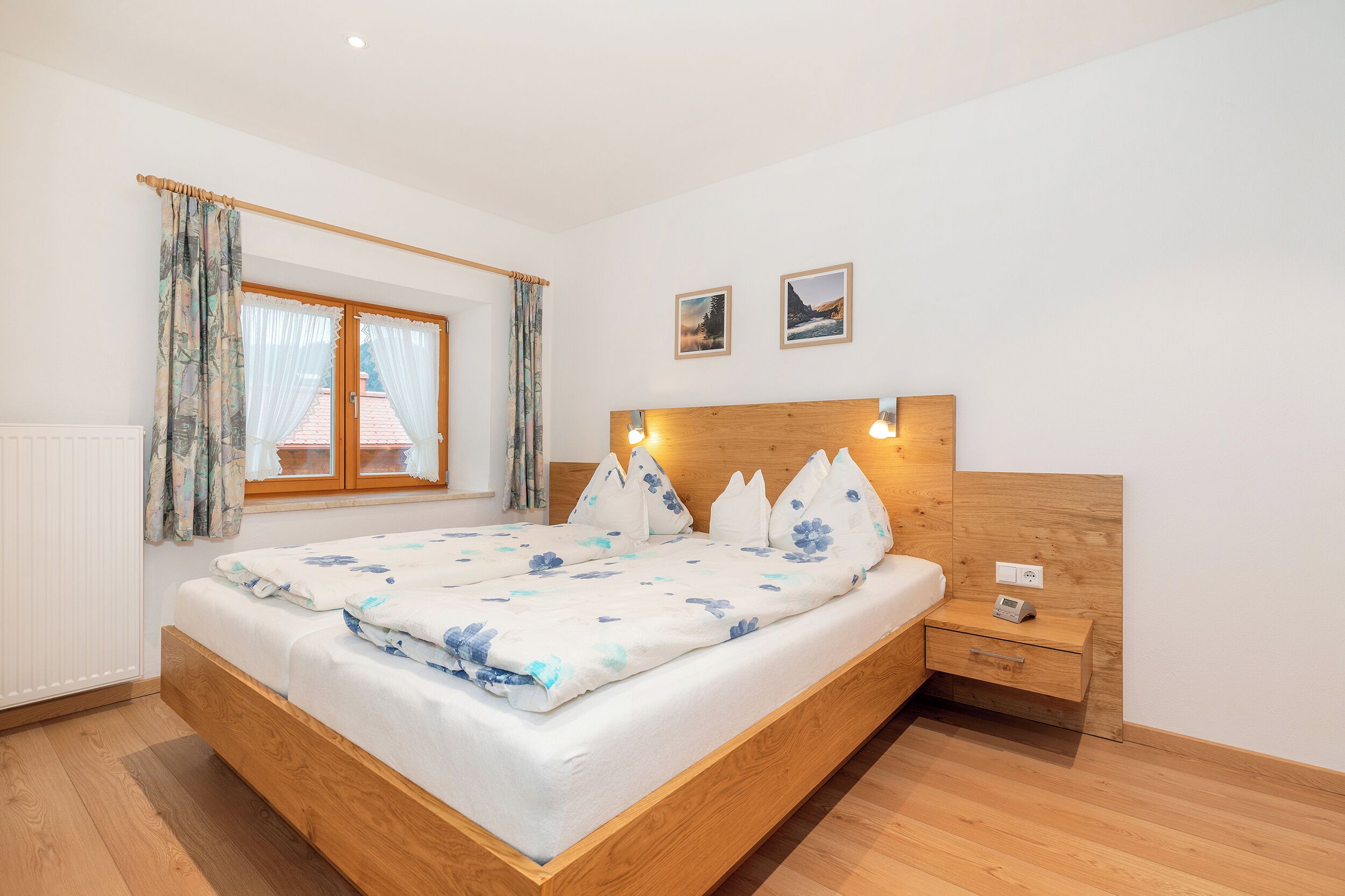 Een slaapkamer met een houten bed, een nachtkastje, een raam met gordijnen en twee ingelijste foto’s aan de muur.