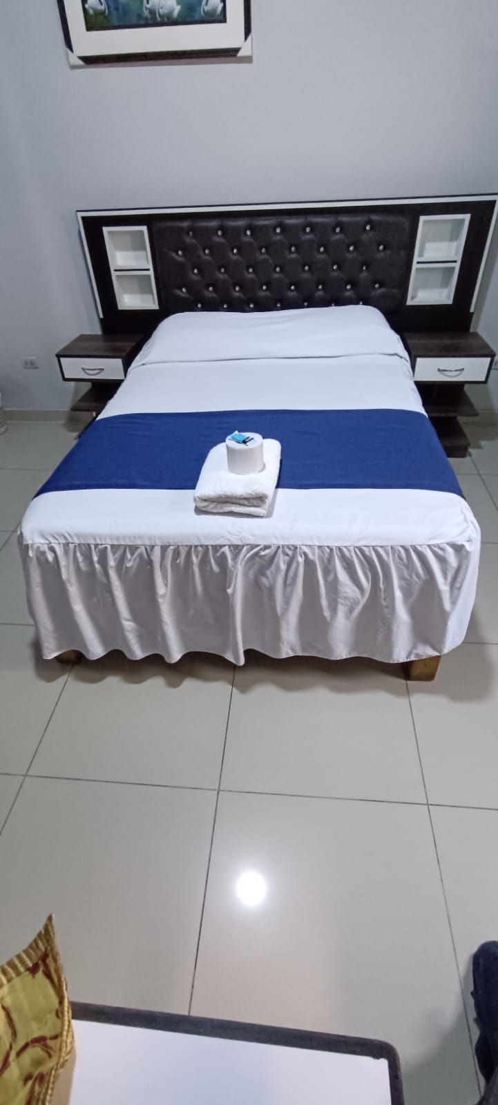 A neatly made bed with a white and blue bedspread, a folded towel, and a dark headboard.