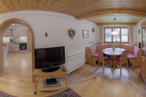 Un salon chaleureux avec un téléviseur, des meubles en bois, un coin repas avec une table et des chaises, et une cuisine en arrière-plan.