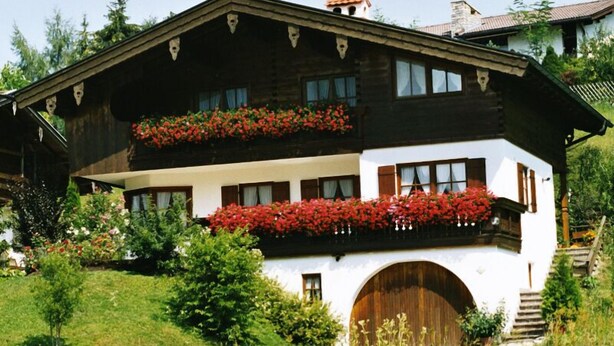 Un chalet traditionnel avec une façade en bois, une partie inférieure peinte en blanc et une grande porte en bois. Il est doté de balcons ornés de fleurs rouges et est entouré d’une végétation luxuriante.