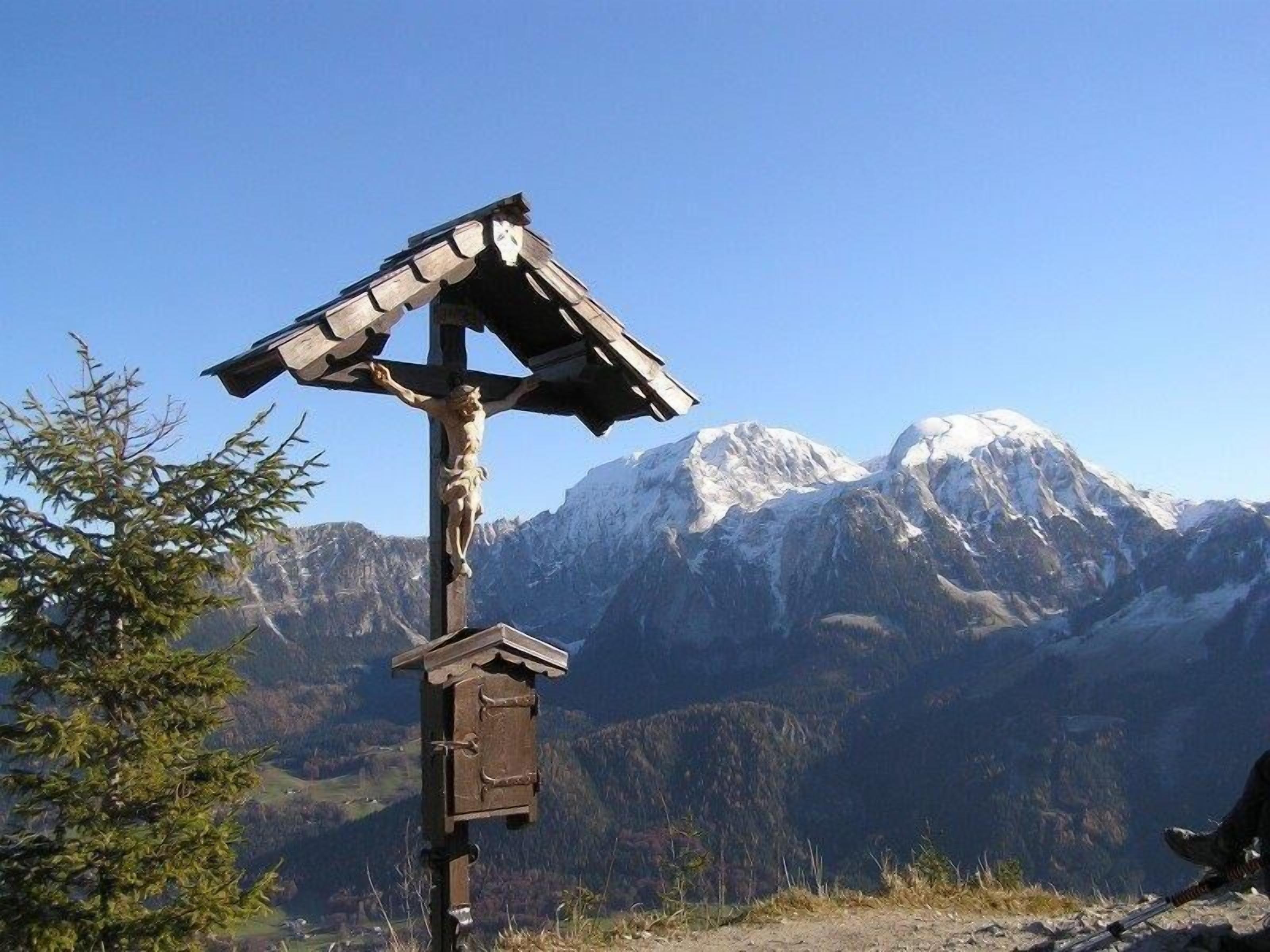 Una cruz de madera con un pequeño refugio para pájaros, con montañas nevadas al fondo.