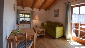 A cozy room with a wooden ceiling, a green sofa, a wooden table with chairs, a lamp, and a window with curtains. - 5. Fewo Hochgern, 37 qm, 1- 3 Pers., 1 Schlafzimmer, 1 Wohnschlafraum, Süd-Balkon (Grassau)