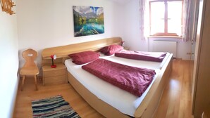 A bedroom with a large bed, a nightstand, a chair, a painting on the wall, and a window with curtains. - Ferienwohnung III° extra Schlafzimmer, Süd-Balkon, 65 qm° (Aschau im Chiemgau)