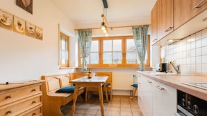 A kitchen with wooden cabinets, a dining table with chairs, a window with curtains, and a tiled floor. - Ferienwohnung III° extra Schlafzimmer, Süd-Balkon, 65 qm° (Aschau im Chiemgau)
