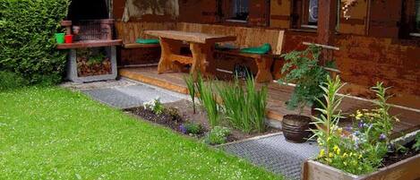 A rustic wooden house with a garden area featuring a wooden bench, a small table, and a raised garden bed with flowers.