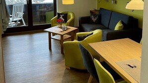 A modern living room with a wooden floor, green and black sofas, a wooden dining table, and a large window with a view of a building outside.