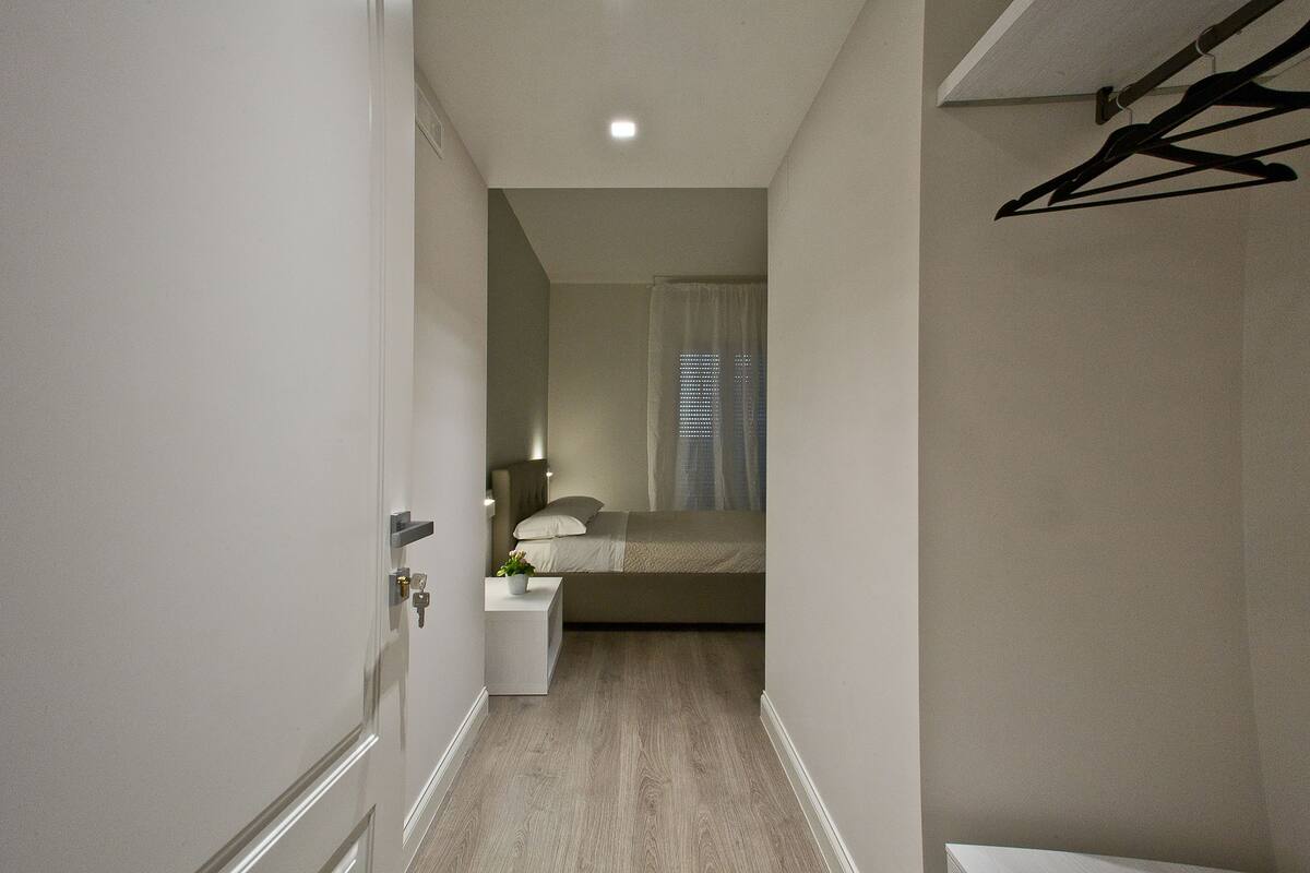 Un couloir étroit mène à une chambre à coucher qui contient un lit, une table de chevet et un porte-manteau mural.
